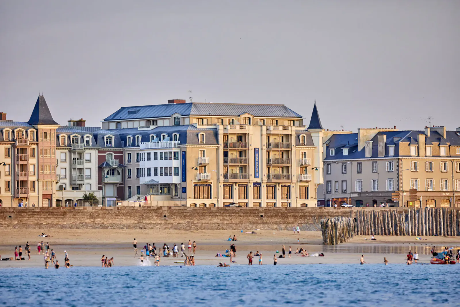 Colorful waterfront apartments in Saint-Malo with beach, people swimming in sea, and pier at sunset.