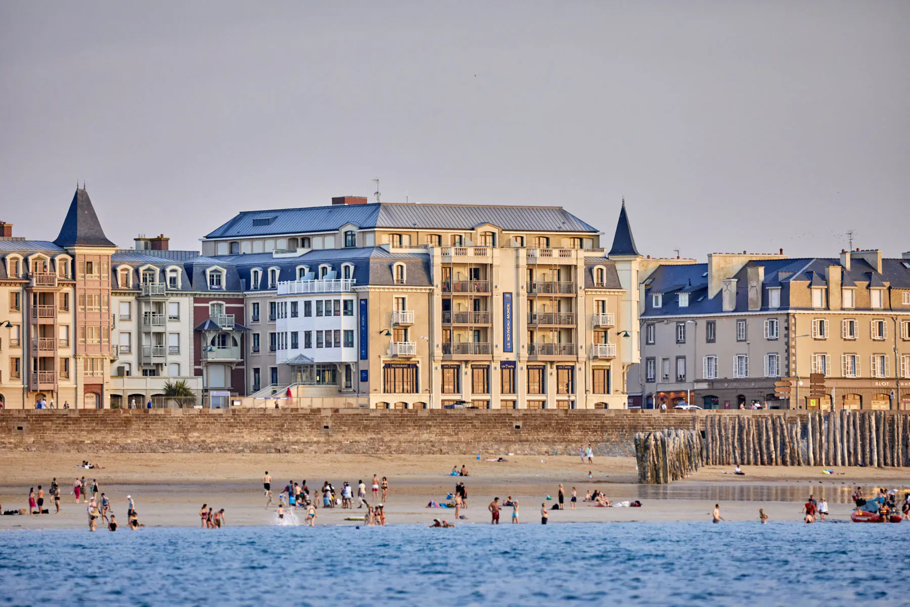 Colorful waterfront apartments in Saint-Malo with beach, people swimming in sea, and pier at sunset.