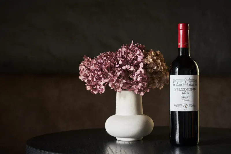 Red Vergenoegd Löw wine bottle beside white vase of pink and beige hydrangeas on black table.