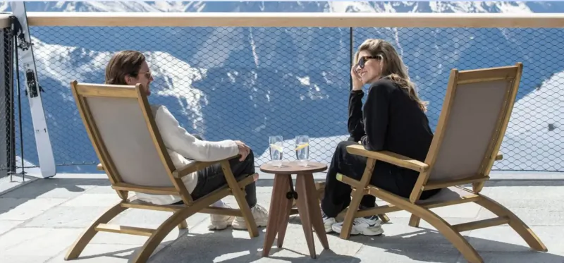 Man and woman in sunglasses sit chatting on wooden chairs with drinks at snowy mountain chalet balcony.