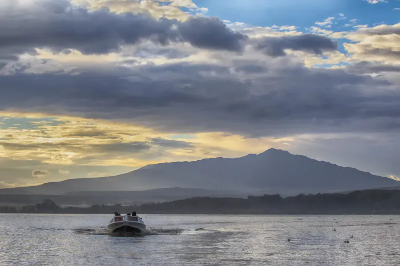 Small motorboat with two people cruising on Lake Naivasha toward mountain at sunset, dramatic clouds, Kenya.