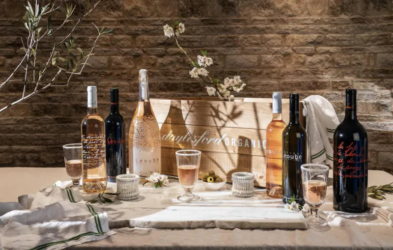 Assortment of wine bottles in wooden crate on table with rosé glasses, olives, and cherry blossoms against brick wall.