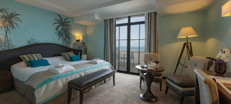 Luxurious hotel room in Saint-Malo with king bed, palm wallpaper, balcony sea view, armchairs, and lamps.