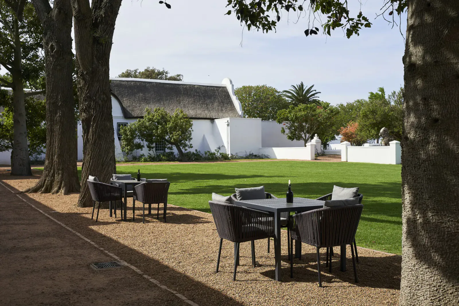 Outdoor patio at Clara’s Barn, Vergenoegd Löw: white thatched building, tall trees, set table with wine bottle on gravel path by green lawn