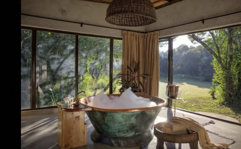 Luxurious copper bathtub with bubbles in eco-lodge bathroom, large windows overlooking Maasai Mara trees and savanna.