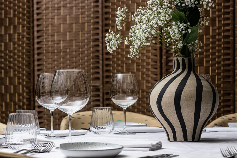 Dining table set with striped black and white vase of baby's breath, wine glasses, plates, and cutlery against bamboo screen.