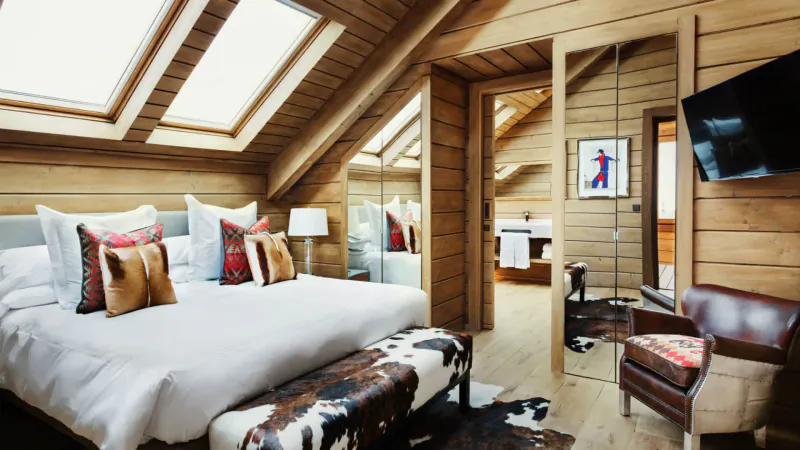 Cozy wooden attic suite at El Lodge, Sierra Nevada, with king bed, colorful pillows, cowhide bench, armchair, and TV.