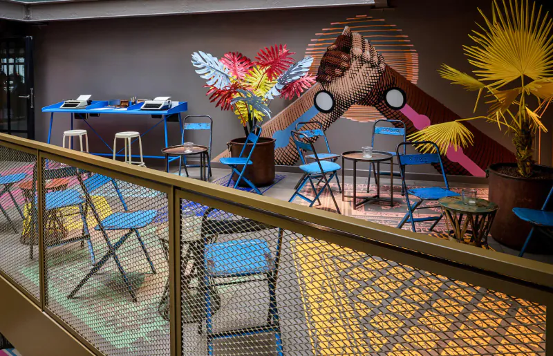 Vibrant 25Hours Paris gallery lounge with blue chairs, tropical plants, and mural of clasped hands on gray wall.