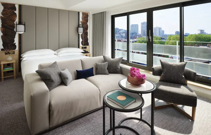 Luxurious Nobu Hotel suite with king bed, plush sofa, coffee tables, pink flowers, and city skyline view from large windows.