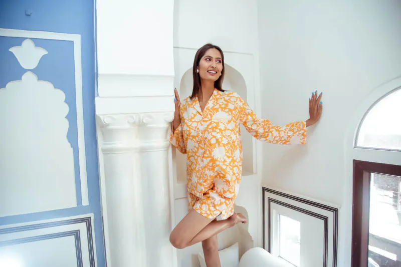 Smiling woman in orange floral long-sleeve kaftan poses playfully with one leg raised in elegant white room with blue arch.