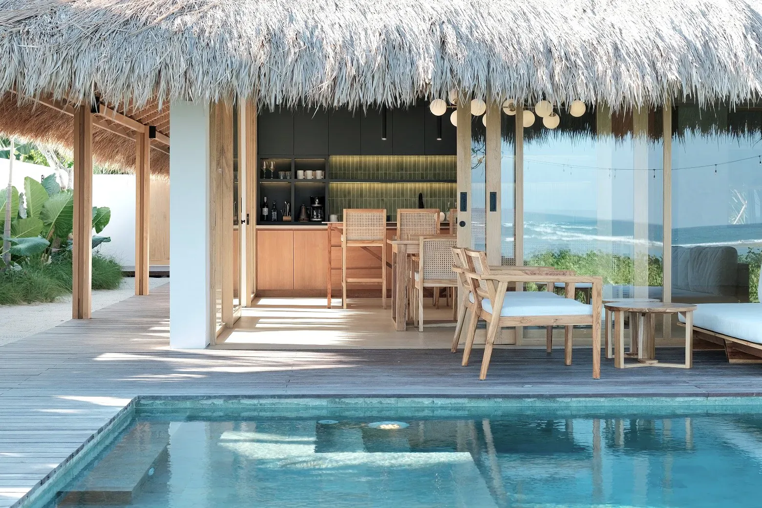 Open-air kitchen and dining area with wooden furniture under thatched roof at beachfront resort, overlooking turquoise pool and ocean.