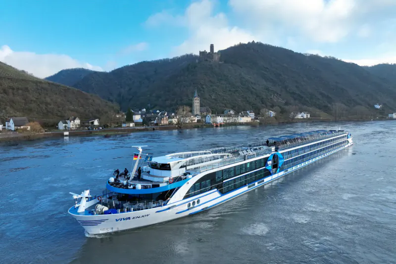White Viva Cruises river ship numbered 185 sails Rhine River past Marksburg Castle on forested hill and village under blue sky.