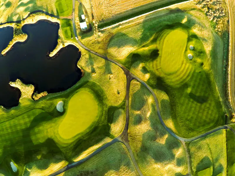 Aerial view of lush green golf course with bunkers, ponds, paths, and surrounding fields.