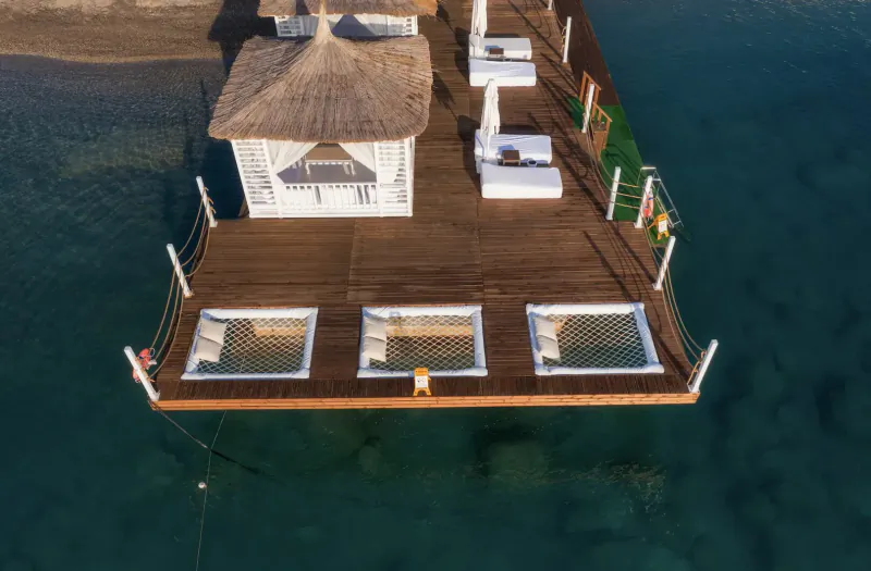 Aerial view of wooden pier over blue water with white thatched cabana, loungers, umbrellas, and netted hot tubs.