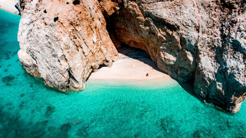 Aerial view of secluded sandy beach nestled between red rock cliffs in turquoise Greek waters, with a tiny figure on shore.
