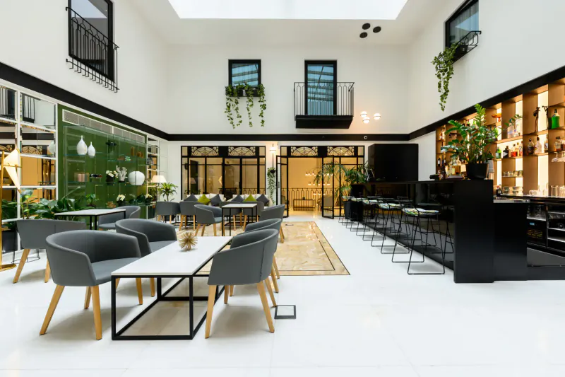Modern luxury hotel atrium in Embassy Valletta with white walls, green plants, wooden dining tables, bar, and skylight.