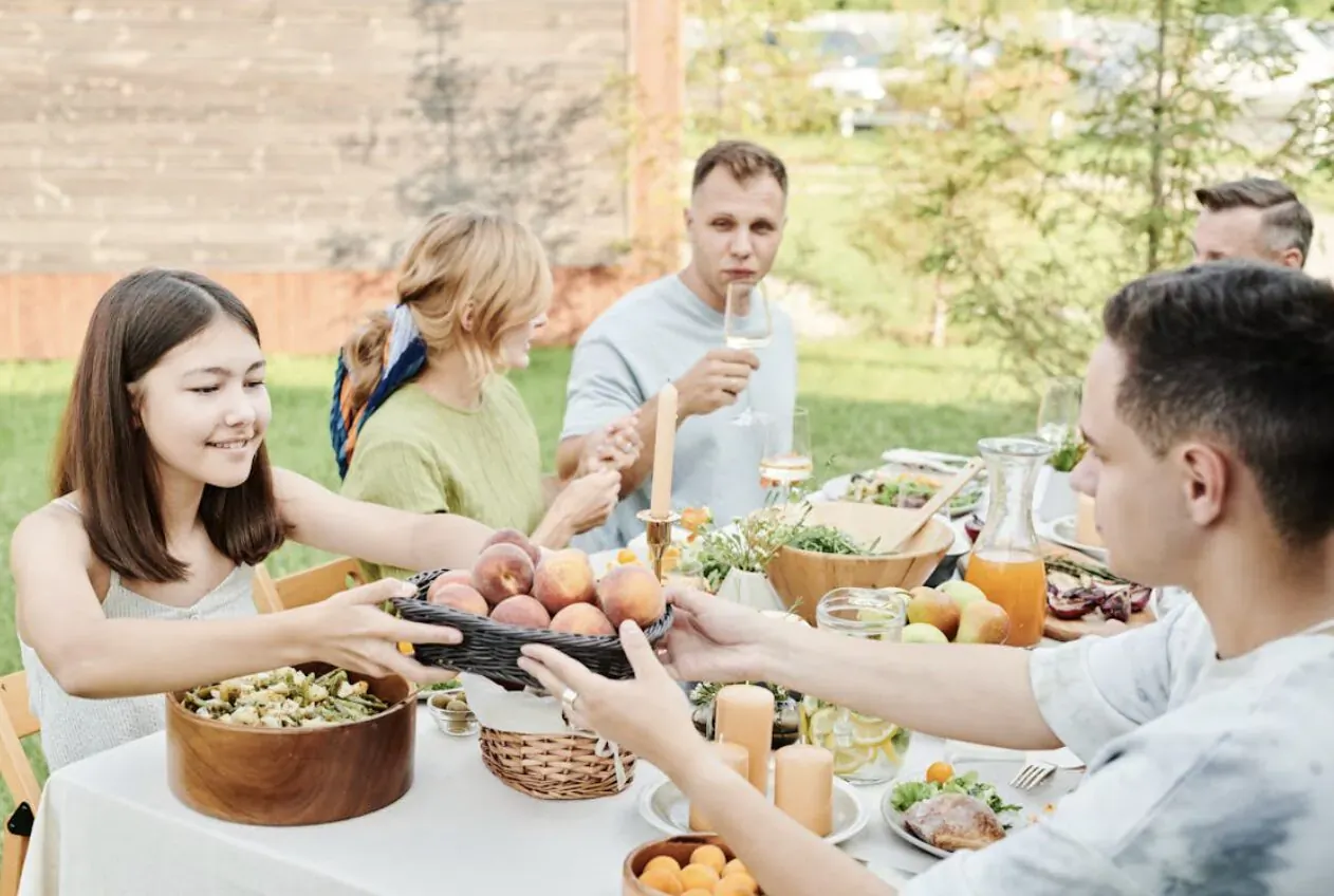 Family passing bowl of peaches at outdoor summer dinner table with food and drinks.