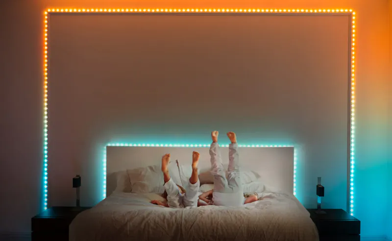 Two children in pajamas playfully standing on head on bed, illuminated by colorful Twinkly Dots LED strips on wall.