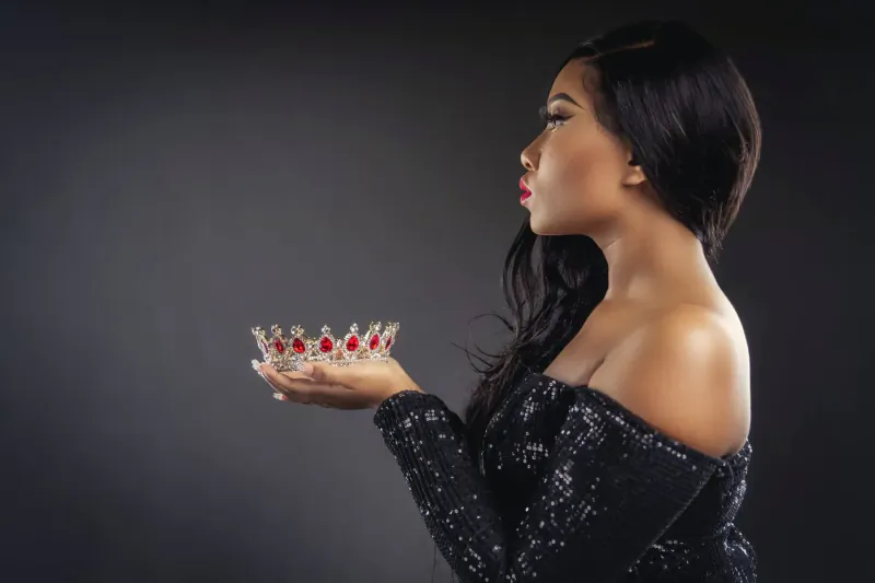 Profile of Asian woman in off-shoulder black gown holding ornate red-gem crown, Regency style, dark background