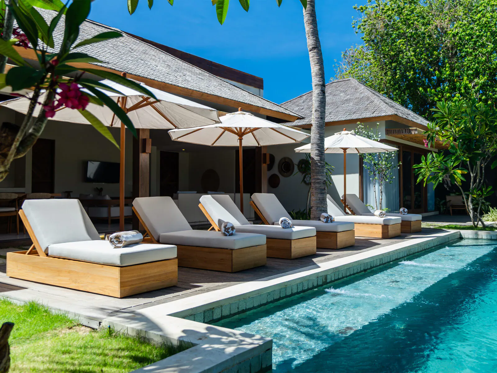 Luxury wooden sun loungers with towels by infinity pool at tropical resort, umbrellas, palms, blue sky.