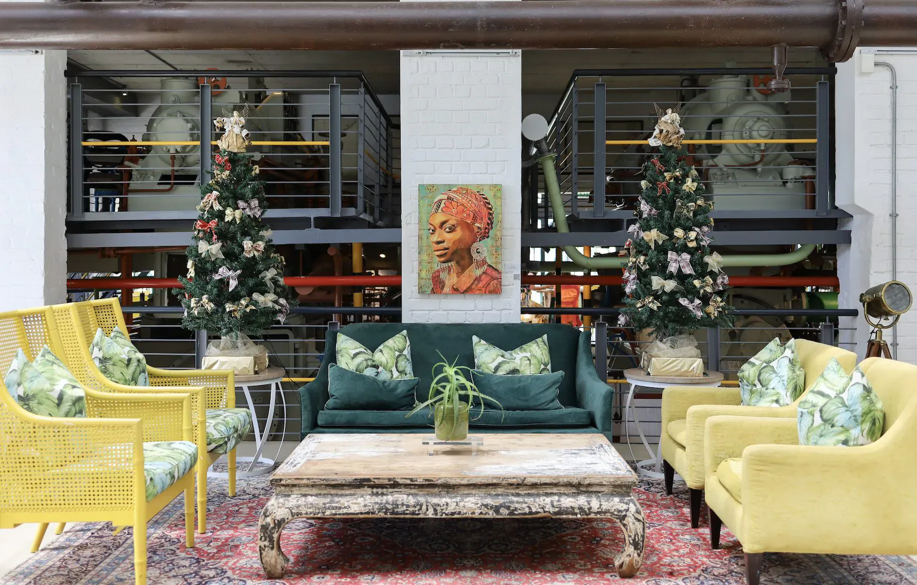 Cozy lounge in Turbine Hotel with yellow chairs, green sofa, rustic table, Christmas trees, and portrait of woman in headwrap against industrial backdrop.