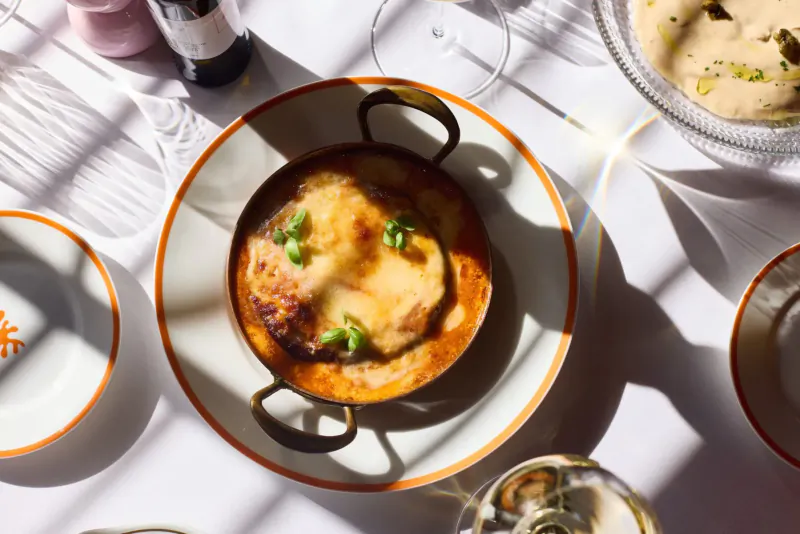 Close-up of cheesy gratin with peas in orange-rimmed cast iron dish on white table with wine glasses, salt, and side dishes at Italian restaurant.