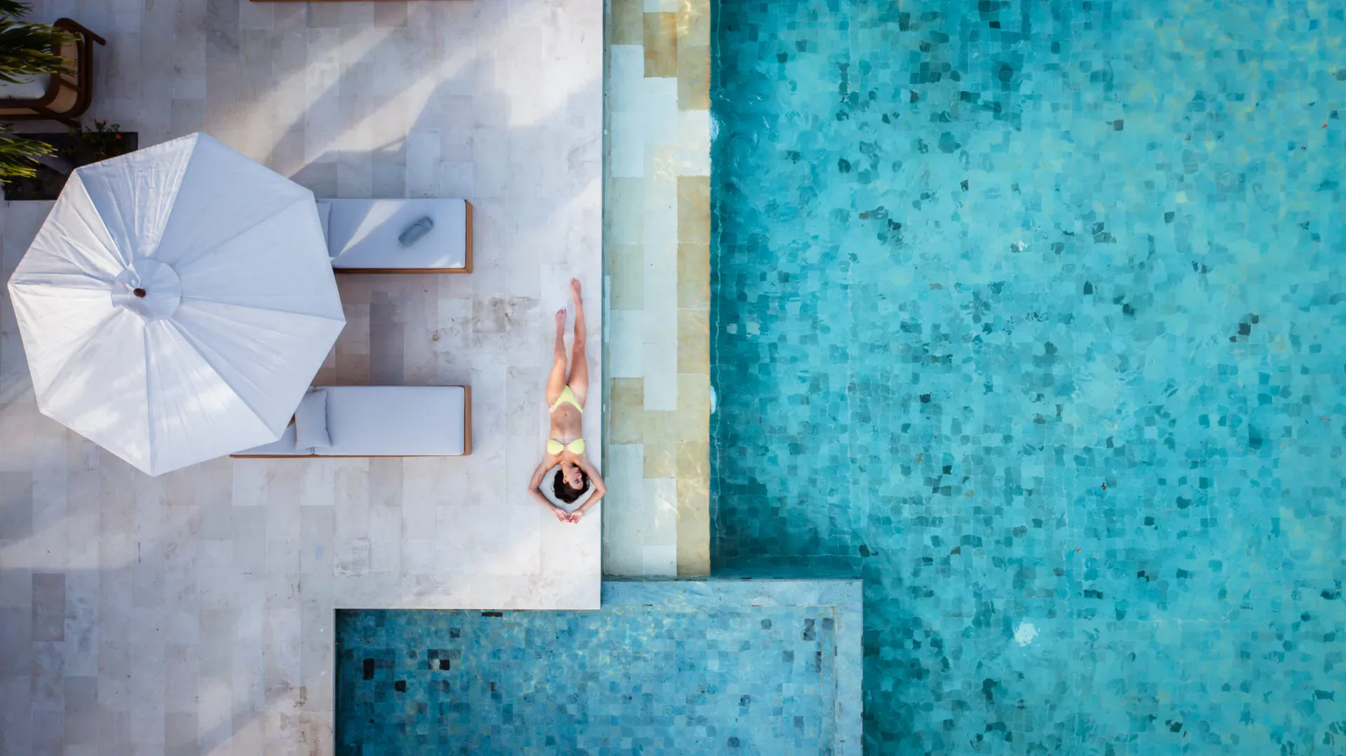 Aerial view of woman lounging by infinity pool at luxury resort, white umbrellas and loungers on tiled deck.