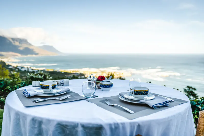 Elegant table set for two with cups and plates on white cloth, overlooking Cape Town's ocean and mountain view.