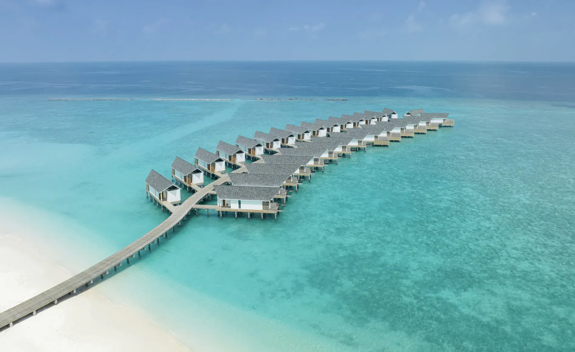 Aerial view of curved row of white overwater villas connected by wooden walkway in turquoise Maldives lagoon