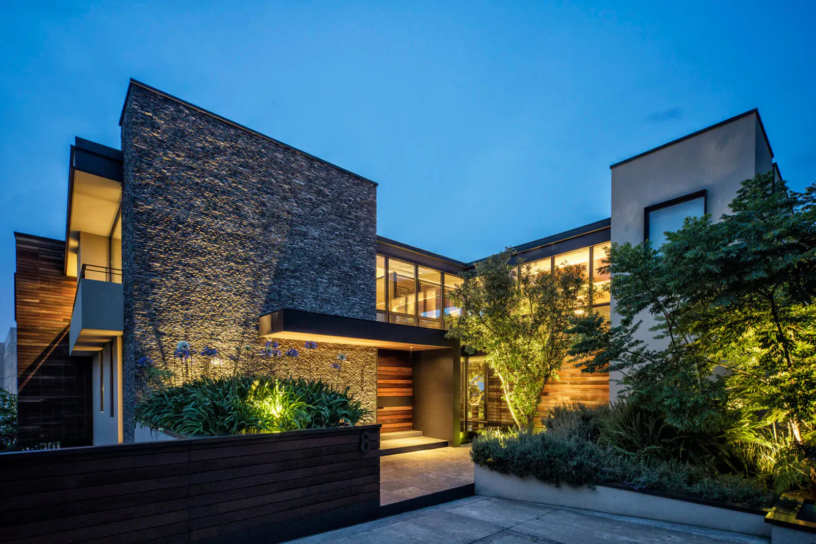Modern Mexican stone and wood house at dusk, lit warmly with landscaped greenery and entry lights.