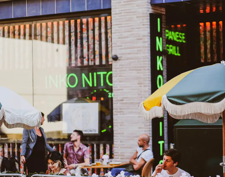 Outdoor Japanese restaurant 'INKO NO ITO' in Carnaby London with neon sign, patrons dining under yellow-green umbrellas.