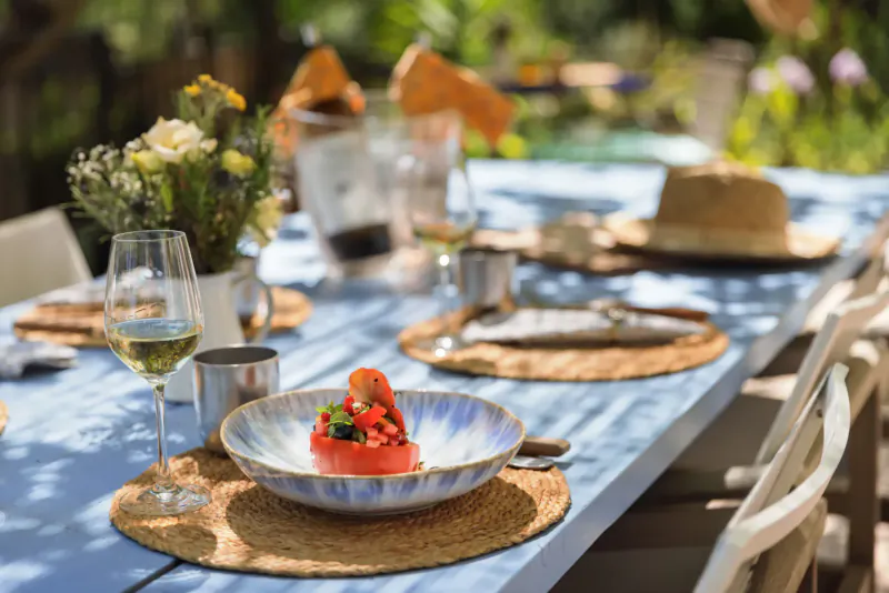Close-up of tomato tart on blue plate with white wine glass on outdoor table at Provence hotel, surrounded by flowers and hat.