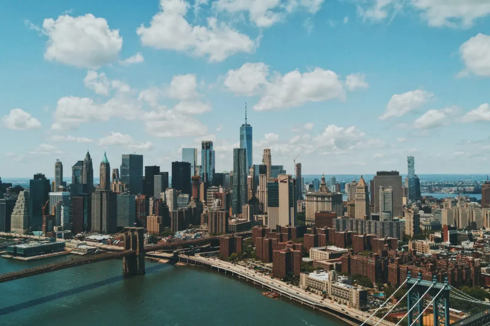 Aerial view of New York City skyline with One World Trade Center and Brooklyn Bridge over the East River under blue sky.