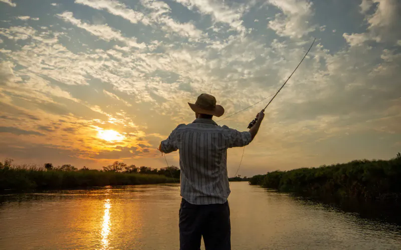Man in straw hat fly fishing from riverbank at sunset in Okavango Panhandle, Botswana