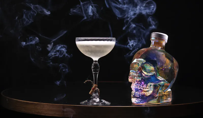 Creamy cocktail in ornate coupe glass with tentacle stem beside iridescent skull bottle on smoky wooden bar table