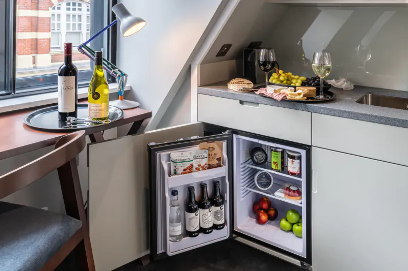 Open mini fridge in cozy London hotel kitchenette with wine bottles, snacks, fruits, and drinks on counter.