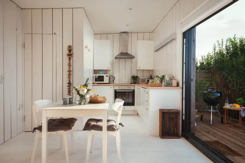 Compact white wooden kitchen-dining area in Sealight clifftop Cornwall cabin, with table, chairs, flowers, herbs, and deck BBQ view.