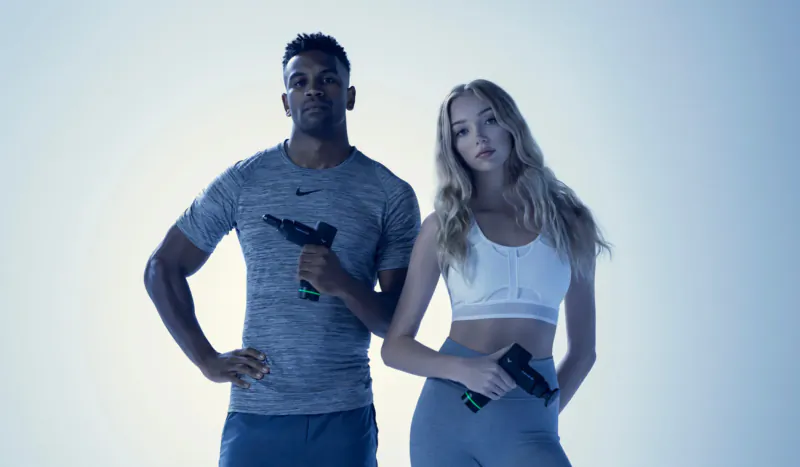 Fit Black man in gray Nike shirt and white woman in white sports bra and leggings hold Hypervolt GO massage guns on blue background