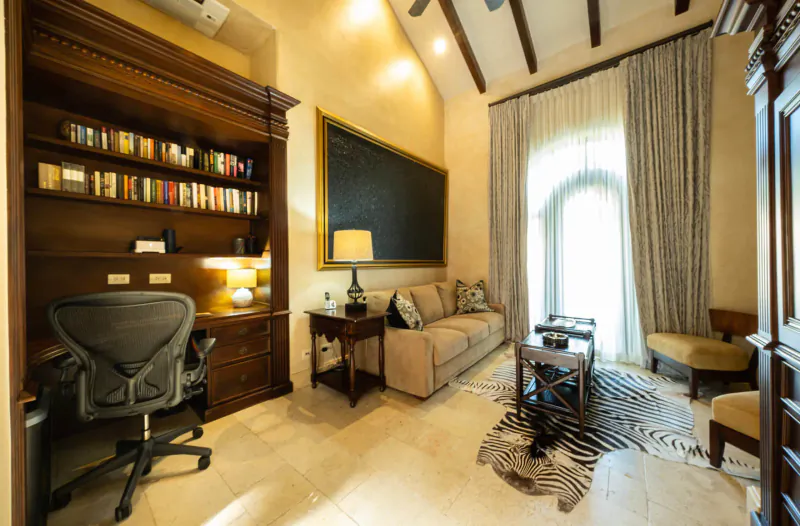 Luxurious villa office with wooden bookshelves, ergonomic desk chair, beige sofa, zebra rug, and arched window.