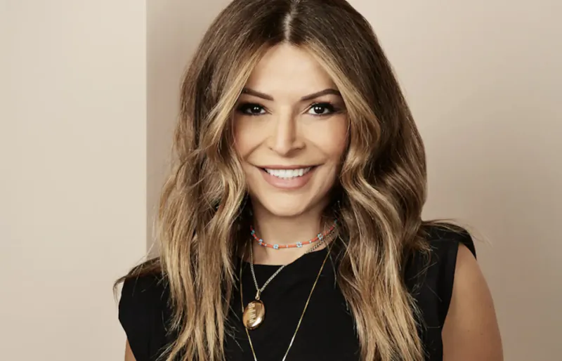 Smiling woman with wavy highlighted hair, wearing layered necklaces and black sleeveless top, in salon gallery