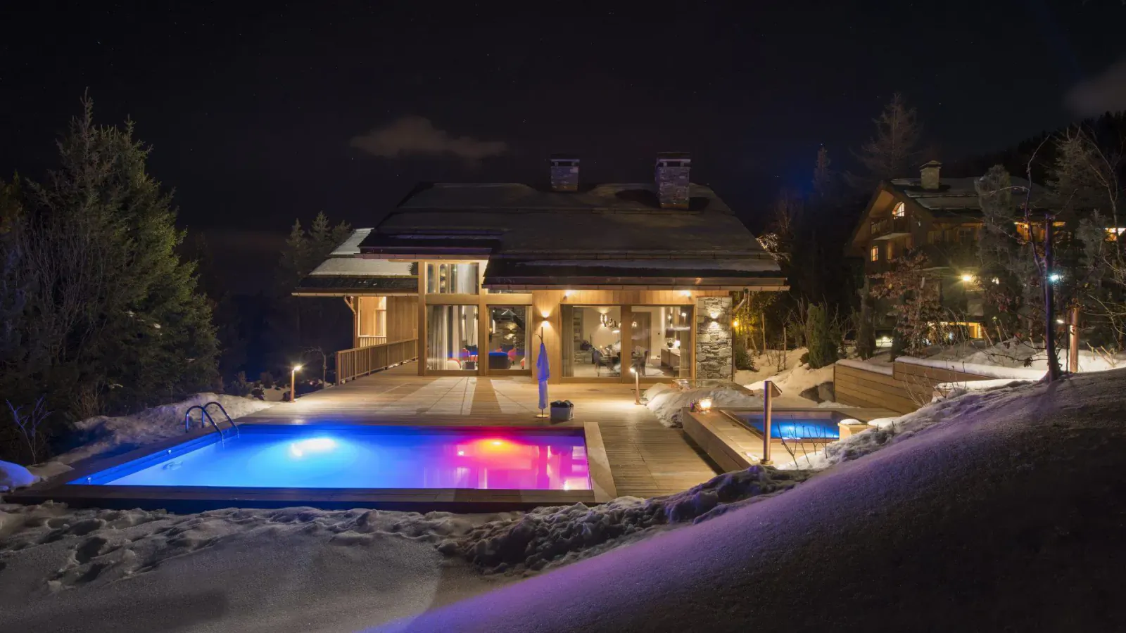 Luxury ski chalet at night with glowing blue-pink infinity pool, hot tub, snow, and pine trees in exclusive resort