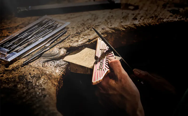 Hands crafting Aston Martin winged logo stamp on paper with tools on rocky surface