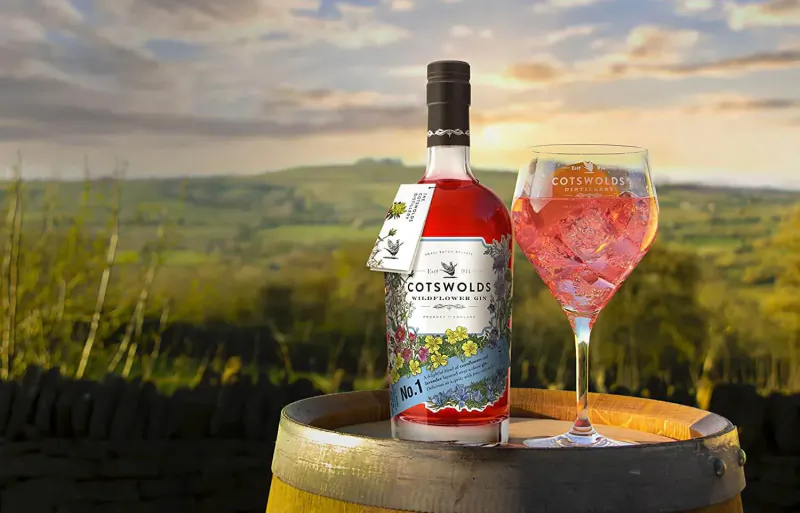 Cotswolds gin bottle with pink gin and tonic in glass on barrel, scenic countryside sunset backdrop