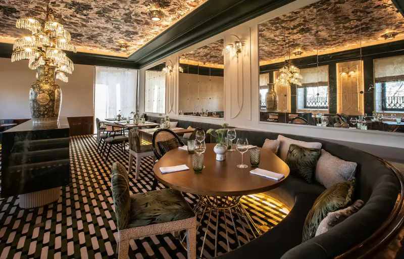 Elegant Vinha Boutique Hotel dining room in Porto with ornate ceilings, chandeliers, round wooden table, green velvet banquettes, and large vases.