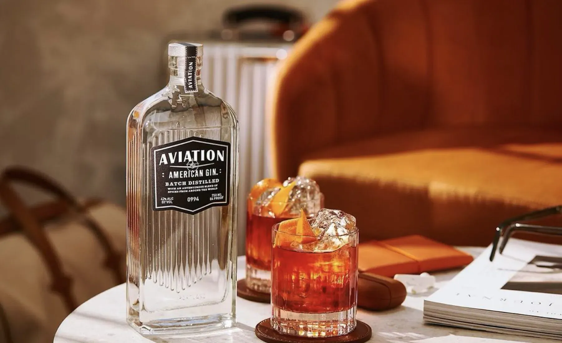 Aviation American Gin bottle beside two orange gin cocktails on a table near orange velvet chair