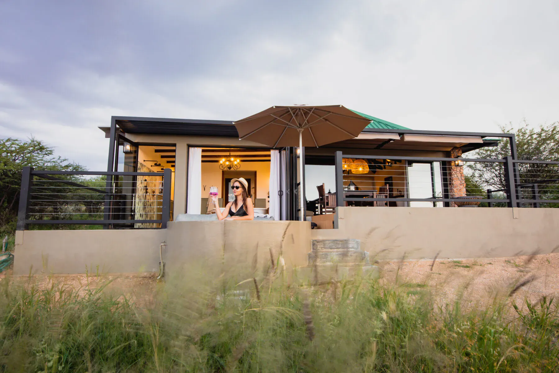 Woman waving from modern Namibia lodge deck with umbrella, surrounded by savanna grass under cloudy sky