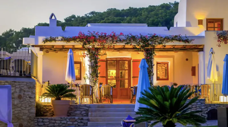 Twilight view of Rural Hotel Can Lluc gallery in Ibiza: white facade with bougainvillea, blue umbrellas, palms, tables, and lanterns.