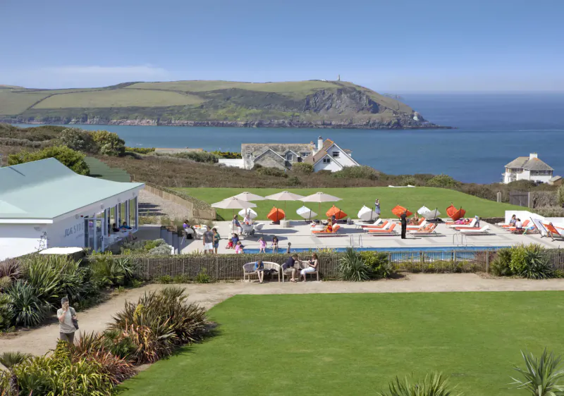 Aerial view of St Moritz Hotel in Cornwall: infinity pool with orange loungers, white building, green lawns, ocean and cliffs beyond.