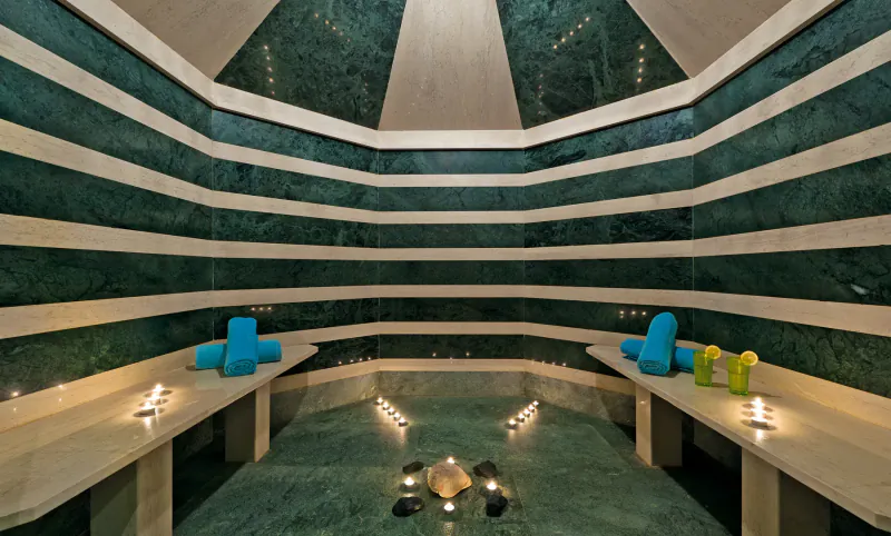 Luxurious octagonal steam room with green marble walls, blue towels on benches, candles, and steaming water in center.