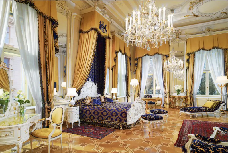 Opulent rococo bedroom in Hotel Imperial Vienna with gold curtains, crystal chandeliers, carved bed, and antique furnishings.
